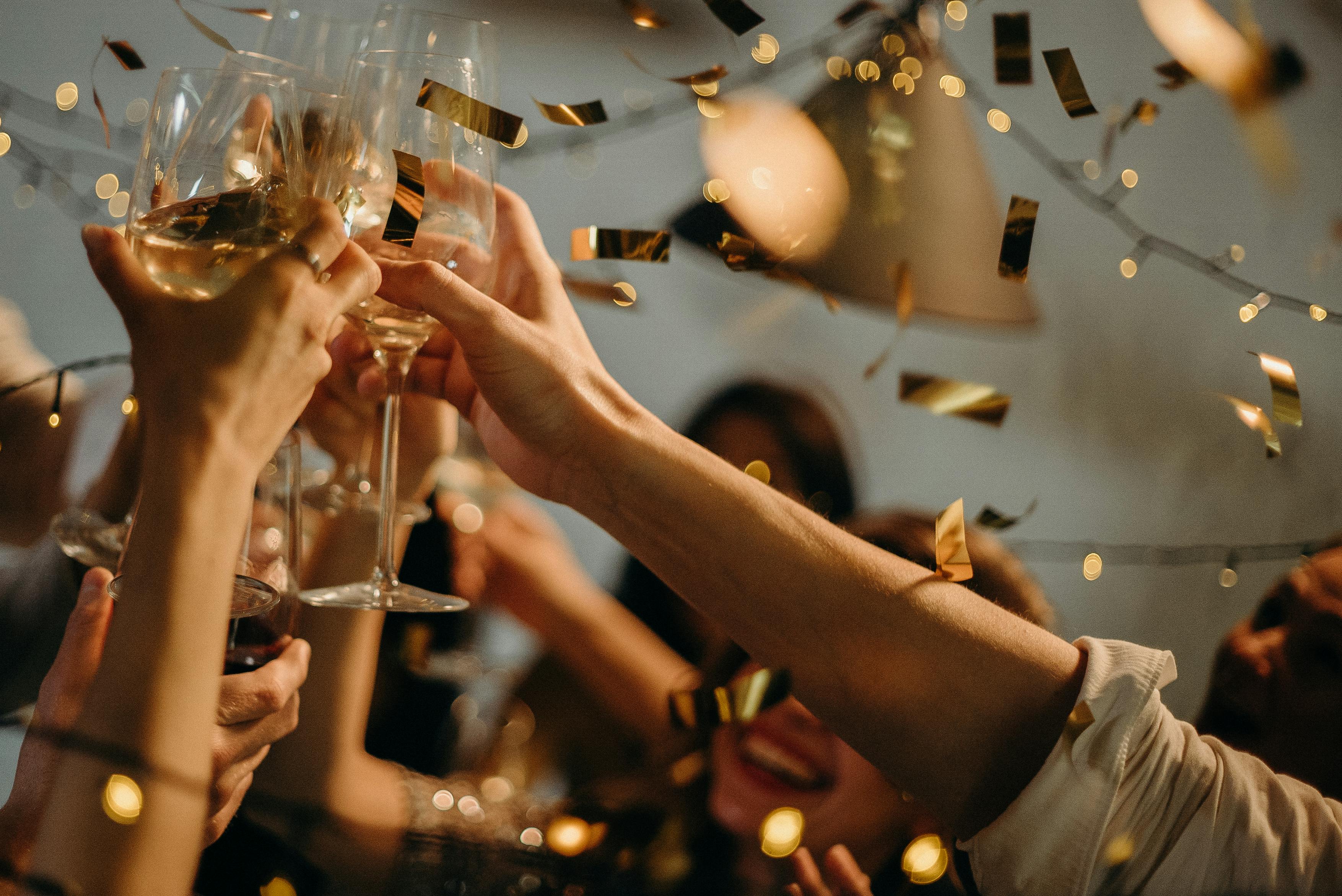 Group of people raising their glasses in a toast at a festive celebration, surrounded by golden confetti and warm lighting.